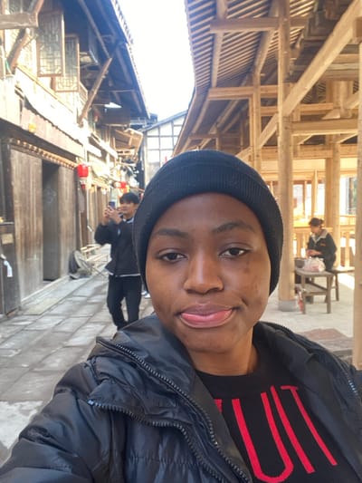 A selfie captures a person wearing a black beanie, a black sweater with red stripes, and a black jacket. They're standing outside, possibly in a market or village. Traditional wooden architecture is visible in the background, including roofs and walkways. Other people can be seen walking or sitting in the background, some appearing to be engaged in daily activities. The lighting suggests it is daytime.