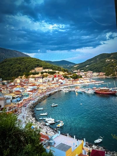 This image presents an elevated, wide-angle view of a densely populated coastal town and its bay, identified as being located in Kumanovo, North Macedonia. The scene is dominated by a clear, turquoise-to-deep-blue bay, bordered by a curving rocky seawall and a narrow paved promenade. Numerous Mediterranean-style buildings with light-colored facades and terracotta roofs are tightly clustered along the waterfront and extend up the lower slopes of steep, heavily vegetated hills that encircle the bay. The sky above is characterized by dramatic, dark blue-grey clouds, with brighter patches near the horizon. The promenade is populated by a significant number of individuals engaged in walking and dining at numerous outdoor establishments marked by umbrellas and canopies. The bay contains a variety of vessels, including one prominent large red multi-decked boat, several medium-sized white yachts, and numerous smaller white recreational or fishing boats anchored or moored. Green foliage partially obscures the immediate foreground on the left.