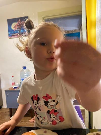 The image shows a young girl with blonde hair, wearing a white t-shirt with Minnie Mouse designs. She is looking at the camera with her hand extended forward, partially obscuring the lens. The girl appears to be in an indoor setting, possibly a home. In the background, there is a large blue cabinet, a bottle of water, and a painting of a tree under a full moon. The overall lighting suggests it is daytime, with some artificial light sources visible. The environment appears somewhat cluttered, consistent with a domestic setting. The location is specified as Mariupol, Ukraine.
