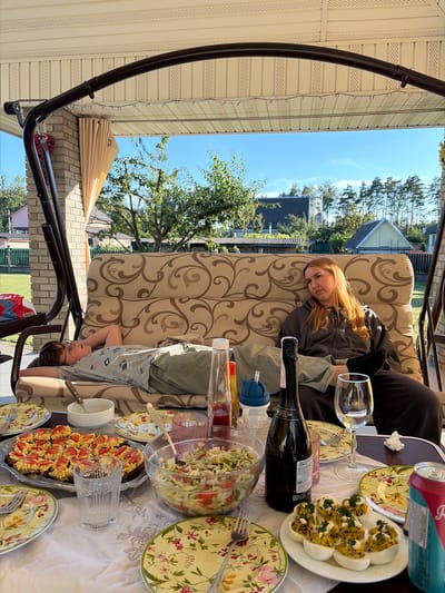 On a sunny day in Навадворскі сельскі Савет, Belarus, a family enjoys a meal on a porch. A boy, wearing glasses and a light-colored t-shirt with a graphic print, lies on a patterned swing set cushion, his hands behind his head. Next to him, a woman with long, reddish-blonde hair sits on the same cushion, looking to her right, wearing a dark hooded jacket and dark pants.
A table is set with various dishes. In the foreground, several plates with a floral pattern are visible. A glass of clear liquid stands near a large metal platter filled with what appear to be open-faced sandwiches topped with red and yellow ingredients. A clear glass bowl contains a salad of chopped vegetables. Several deviled eggs are arranged on a white plate, garnished with green herbs and yellow seasoning. A bottle of sparkling wine and a wine glass are also on the table, along with a can of soda and a bottle of ketchup.
The porch has a brick pillar and a beige ceiling with decorative trim. Beyond the porch, a green lawn is visible, with trees and houses in the background. The sky is clear and blue. The overall atmosphere suggests a relaxed outdoor gathering.