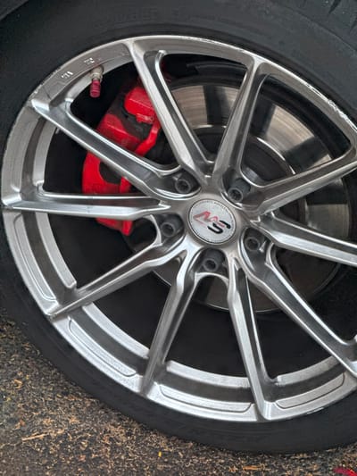 This media file is a close-up, medium-shot photograph of a car wheel. The wheel is silver with a multi-spoke design. The tire is black and appears to be in good condition. Visible behind the wheel is a red brake caliper and the brake rotor. There is a circular sticker in the center of the wheel that appears to have red and black lettering and a logo. Some of the text on the sticker is visible and reads: "PERFORMANCE WHEELS BY SPACWHEELS" and "RS". The wheel has some minor scuff marks and scratches, particularly around the outer rim. The setting appears to be outdoors, with what looks like a textured ground surface with small debris visible to the right of the wheel. The lighting suggests it is daytime. The image is static and does not depict any activity or event taking place. There are no people visible in the image. The location context is Adeje, Spain.