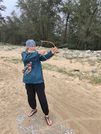 An adult female, appearing Caucasian, is depicted standing on a sandy expanse identified as Thuận An Ward, Vietnam, under an overcast sky. She is oriented slightly right of center, aiming a primitive bow and arrow directly forward.

The subject wears a blue and white patterned beanie, a teal lightweight jacket, black casual pants, and black thong-style sandals. A colorful patterned fanny pack is secured across her chest. Her blonde hair is braided and visible beneath the beanie. Her hands manipulate a reddish-brown curved stick bow with a white string, and a thin, light-colored stick serves as an arrow, drawn back for release.

The immediate ground is sand with sparse green vegetation. Scattered grey rocks and concrete debris are present in the midground to the right, along with incidental litter. A dense line of tall, slender evergreen trees forms the backdrop. Faint tire tracks are visible in the foreground sand.