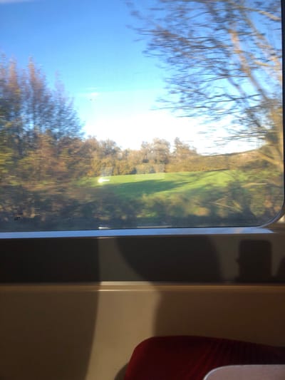 The image captures a dynamic view from inside a moving train, depicting the landscape in the vicinity of Dettwiller, France. The upper frame is dominated by a clear, bright blue sky, with the sun causing a localized glare near the horizon. The exterior scenery is rendered with horizontal motion blur, consistent with a moving vehicle. On the left, a dense cluster of deciduous trees displays blurred brown and orange foliage. To the right, a bare deciduous tree with prominent branches is similarly blurred. The mid-ground features a vibrant green grassy field that slopes gently upwards, with faint shadows indicating undulations. A distinct, bright white rectangular object rests on the green field, positioned slightly left of the image center. The lower portion of the image reveals the interior of the train carriage: the lower frame of a window, which includes a small white "AGC" label in its bottom-left corner, rests atop a light-colored horizontal panel. Below this, a darker gray panel is visible. The top right segment of a red upholstered train seat is present in the bottom right corner. A prominent, dark shadow resembling a human head and upper torso is cast onto the darker gray interior paneling, suggesting the reflection of a person, likely the photographer, inside the train.