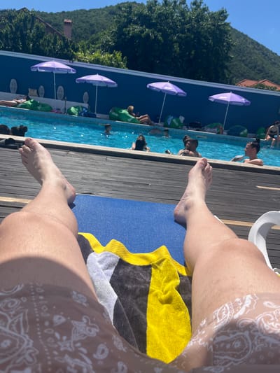 The image depicts a perspective shot of someone relaxing on a lounge chair by a swimming pool. The person's legs and feet are prominently in the foreground, with a towel draped over the chair. In the background, a blue-tiled swimming pool is filled with people swimming and lounging. Purple umbrellas provide shade around the pool area, and beanbag chairs are scattered on the deck. The scene is set outdoors on a sunny day, with lush green trees and a hillside visible in the distance. The overall atmosphere suggests a resort or a recreational area. This location is likely a pool area within the town of Varvara, Bulgaria, given the context of the prompt.