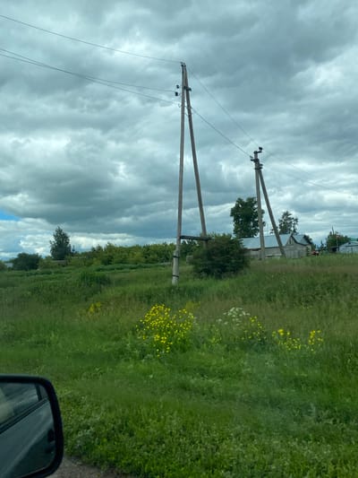 The image depicts a rural landscape in Krasnovka, Russia. The scene is dominated by a dramatically cloudy sky, with dark, heavy clouds suggesting impending rain or a storm. Two wooden utility poles stand prominently in the foreground, stretching upwards towards the overcast sky. Wires are strung between them, a common sight in rural power infrastructure.

To the right of the poles, a lush green field unfolds, dotted with small yellow wildflowers. The field appears to be a roadside verge or a meadow. In the background, a few trees and what appear to be modest houses or farm buildings are visible, peeking through the foliage and under the dramatic sky. The overall impression is one of a slightly somber, natural setting. The image appears to have been taken from a vehicle, as the corner of a black side-view mirror is visible in the lower right of the frame, reflecting the sky and the green landscape. The time of day is unclear due to the heavy cloud cover, but it suggests daytime. No people are visible in the image. There is no visible text.