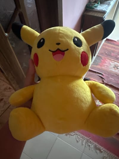 A yellow plush Pikachu toy is held by a hand in the foreground. The toy is facing forward and has red cheeks, black eyes, and a smiling mouth. The background shows a wooden cabinet and a patterned rug. The image was taken in a residential setting.