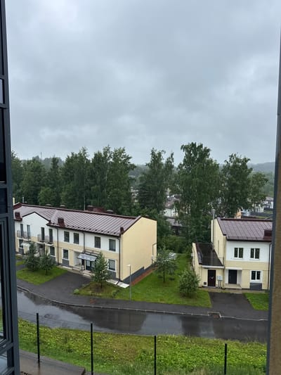 The image captures an outdoor scene of residential buildings on a cloudy, overcast day, likely during daylight hours, given the illumination. The buildings are multi-story, with light-colored facades and dark-colored, sloped roofs. They appear to be modern apartment blocks with multiple windows and balconies. Some buildings have dark doors leading into them, and one has an awning over the entrance.
The scene includes well-maintained green spaces with grass and several small trees planted around the buildings. A paved pathway or road winds through the complex, appearing wet, suggesting recent rain or ongoing precipitation. The presence of a wire fence in the foreground on the right side of the image indicates a boundary or enclosure. In the background, a dense line of trees covers a hill, creating a natural backdrop to the residential development. The overall atmosphere is muted and somber, consistent with the overcast weather. There is no visible text in the image. The location context provided is "Близкое Кп, Russia," which suggests this is a residential area in or near Близкое, Russia.