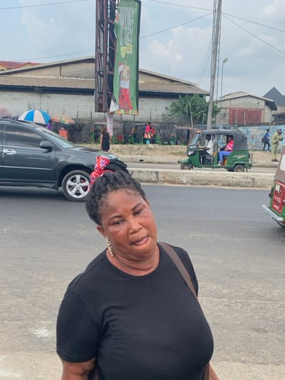 The image depicts a busy street scene in Aba, Abia, Nigeria, under an overcast sky. In the foreground, an adult female with dark skin and black braided hair, accented with a red and white checkered bow or fabric, faces slightly left of the camera. She wears a plain black short-sleeved top and a brown bag strap over her right shoulder. Gold-colored dangling earrings are visible. Her mouth is slightly open, revealing a portion of her tongue, and her facial expression is neutral.
Behind her, a dark-colored SUV is parked on the left side of a paved road, partially obscuring a market area with two vendor umbrellas (one blue, one multi-colored) and several people. A concrete median separates traffic lanes. On the opposite side of the median, a green Keke Napep (auto-rickshaw) moves right to left, occupied by a male driver in a white shirt and a female passenger in pink. Another Keke Napep is partially visible on the far right.
The background features various low-rise buildings with corrugated metal roofs, indicative of an urban or commercial district. A large billboard structure on the left displays a green banner with white text, partially legible as "HAPPINESS IS..." and an image of a red stocking. Utility poles and wires are visible against the light grey sky. Several pedestrians, including a person in a beige uniform, are visible walking in the midground and background.