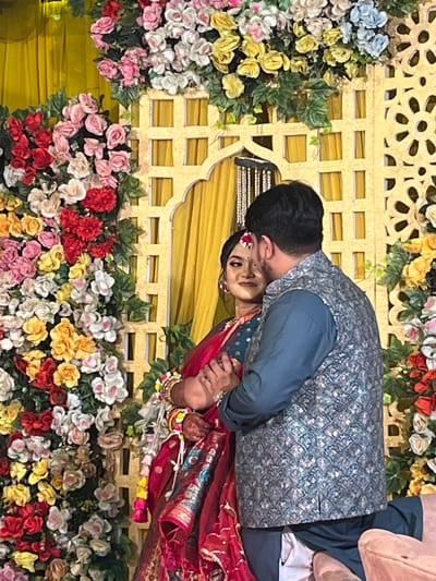 The image depicts two individuals, a male and a female, positioned in front of an ornate floral and lattice backdrop within the city of Savar, Bangladesh. The backdrop features a golden-yellow latticework structure, including an archway and decorative cutouts, heavily adorned with a dense arrangement of artificial flowers in shades of red, pink, white, yellow, orange, and light blue, interspersed with green foliage. Yellow pleated curtains are visible behind the lattice.
The female, positioned on the left and partially facing the viewer, wears a red and dark teal traditional garment, multiple gold bangles on her right wrist, and a red and white floral adornment in her hair. Her face is made up, and she displays a slight smile while looking towards the camera. Her right hand is interlocked with the hands of the male.
The male, positioned on the right with his back predominantly to the camera, wears a dark teal long-sleeved shirt, a patterned grey and silver waistcoat, and dark teal trousers. His hands are clasped with the female's.
A light-colored upholstered armrest is visible in the bottom right foreground.