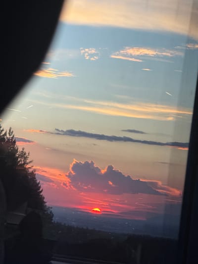 The image depicts a vibrant sunset over a landscape, likely from the vicinity of Baliovtsi, Bulgaria. The sky is a canvas of warm hues, transitioning from deep blues at the top to fiery oranges and reds closer to the horizon. Wispy clouds are illuminated by the setting sun, creating a dramatic contrast. In the foreground, the silhouette of trees lines the left side of the frame, their dark forms stark against the colorful sky. Below the sunset, a distant landscape is visible, appearing as a hazy expanse of muted blues and purples, suggesting hills or mountains. The sun itself is a bright, intense red orb, partially obscured by clouds and the horizon. There are no discernible people or specific identifiable structures within the scene, making it a purely natural landscape captured at dusk.