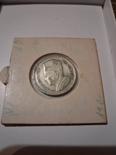 A close-up showcases a silver coin, likely of Middle Eastern origin based on its script, set within a square, light brown cardboard holder. The coin depicts a profile portrait surrounded by Arabic text. Green markings are visible on the cardboard, including the year "1967" and other characters. The coin and holder are situated inside what appears to be a white cardboard box.