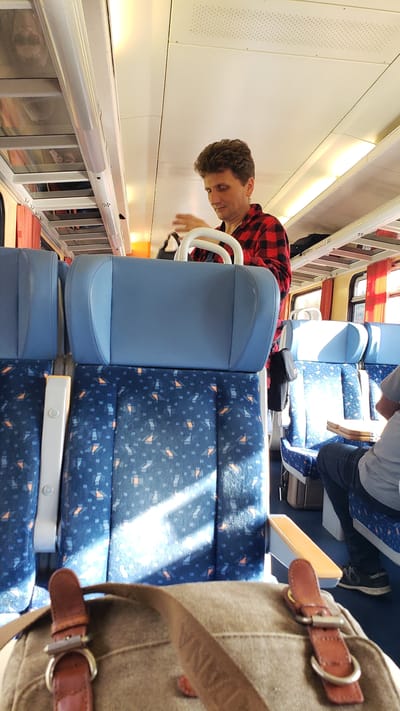 A man with curly brown hair wearing a red and black checkered shirt is standing on a train and adjusting a black bag. He is in the foreground, with the seats of the train in front of him and the ceiling of the train above. The seats are blue with a blue and orange pattern. The train is well-lit with natural light coming through the windows. The man appears to be in the process of stowing or retrieving an item from his bag. The scene is likely within a train carriage, which could be travelling through or stopping at the town of Štrba, Slovakia, as the image metadata suggests.