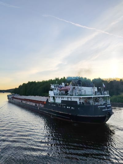 A long cargo ship, identified as "АРКАДИЙ МИНЧЕН" (ARKADY MINCHEN) from its home port of "НИЖНИЙ НОВГОРОД" (NIZHNIY NOVGOROD), navigates a wide river or canal at Volokov Most, Russia. The vessel has a dark hull and a white superstructure at its stern, from which light smoke or exhaust emanates. A Russian flag is visible near the stern. The ship appears to be loaded with bulk cargo, covered by white tarpaulins or covers across its numerous holds.
The scene is captured during either late afternoon or early evening, as indicated by the warm, low sunlight casting a golden glow on the distant treeline and reflected faintly on the water. The sky is a soft blue with subtle wisps of clouds and a faint contrail. The ship moves from the upper left towards the lower right of the frame, leaving a clear wake behind it. The water is calm, reflecting the sky and the ship's dark hull. A dense, dark green forest lines the right bank of the waterway. Another, smaller boat is barely visible in the distance further along the river. The overall atmosphere is peaceful and industrious.