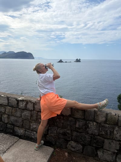 A person in a white t-shirt and orange shorts is standing on a stone wall near the sea in Golubovići, Montenegro. The person is holding a metallic object, possibly a drinking flask, to their mouth. The background includes the sea, sky, and a mountain range. The scene is outdoors during the daytime.