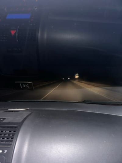 The image captures a nighttime scene from inside a vehicle in Gorna Mahala, Bulgaria, looking forward through the windshield onto a road. The lower third of the frame shows the vehicle's dark gray dashboard, featuring air vents on the left, a blue digital display screen, and a prominent red triangular hazard light button on the central console. Above the dashboard, an object with white markings or text on a dark background hangs from what appears to be the rearview mirror assembly.

The external environment is a dark, unlit road, illuminated by the vehicle's headlights. The road surface, likely asphalt, shows a solid white lane marking on the left and a dashed white lane marking on the right, indicating a two-lane configuration. In the mid-distance on the right side of the road, a blurry, reflective object, consistent with a road sign or marker, is visible. Further ahead, several faint, indistinct lights are discernible in the distance, suggesting other vehicles or distant structures. The sky is completely dark, with no visible features. No individuals are present or discernable within the captured scene.
