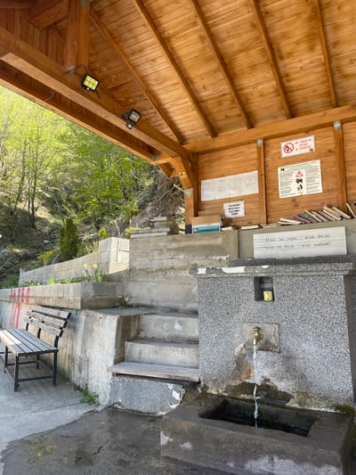 This image depicts a water fountain area, possibly a spring or well, located outdoors. It is likely in the mountainous terrain near Borinovo, Bulgaria, considering the textual information displayed.
The fountain is constructed of gray stone or concrete, with a metal faucet dispensing water into a basin. A set of steps leads up to the fountain, and a wooden bench is situated nearby. The stone wall adjacent to the fountain is inscribed with text in Bulgarian. Additional signage is posted on the adjacent wooden structure, including safety warnings.
The area surrounding the fountain is somewhat wooded. There is an open-air wooden structure above the fountain that appears to provide shelter. Two solar powered lamps are attached to the wooden rafters.