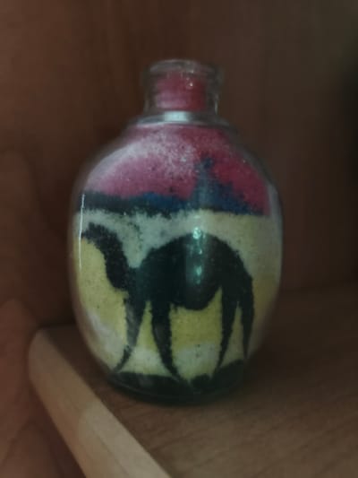A clear glass bottle filled with colorful sand art is displayed on a wooden surface. The sand art depicts a desert scene with a camel silhouette against a backdrop of layered sand in shades of red, blue, yellow, and white. The bottle has a narrow neck with a silver rim. The wooden surface appears to be a shelf or table, with another wooden surface visible above and behind the bottle, suggesting it is placed on a shelf in a cabinet or display unit. The background is dark and out of focus, with a warm brown tone. There are no people or discernible actions depicted in the image. The scene is static and focuses solely on the object. The location of this image is unknown.