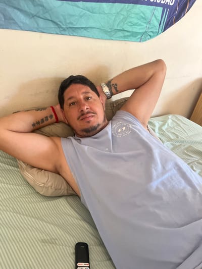 A male subject, appearing to be middle-aged, is depicted lying on his back on a bed in a room in Bernal Este, Argentina. His head rests on a light beige pillow, and his hands are clasped behind his head. He wears a light blue sleeveless top featuring a circular white logo on the left chest. Visible on his left forearm are vertical Asian character tattoos and a red string bracelet. His right forearm displays a script tattoo reading "Samira" and a silver-colored wristwatch. The subject has dark hair and stubble on his chin, looking directly forward. The bed is covered with a green and white striped sheet. To the lower left of the subject, on the bed, is a black remote control with visible app icons (YouTube, Netflix, Disney+, Prime Video). Behind the subject, a light-colored wall is visible, with a blue and teal banner or flag partially suspended, bearing the partially visible text "LA CIUDAD". A wooden furniture item is partially visible on the far right.