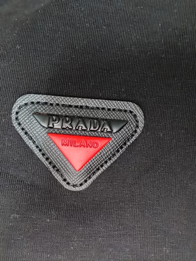 The image presents a detailed close-up of a black textile surface, characterized by a fine, diagonal ribbed texture. Centered in the lower half of the frame is an inverted triangular patch, constructed from a textured black material with a visible black stitched border. The precise dimensions of the patch are not determinable without a reference scale. The word "PRADA" is embossed in raised black capital letters across the upper section of this patch. Directly below, an inverted red triangular insert features the raised white capital letters "MILANO". No individuals, other objects, or actions are depicted. The visual evidence does not provide information to determine a specific location within the city of Saint-Étienne, France.