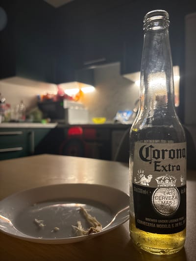 The image depicts a close-up of a partially consumed bottle of Corona Extra beer, placed on a wooden table. A white plate with remnants of food is visible in the foreground. The background shows a kitchen environment, with cabinets, countertops, and illuminated overhead lights. The lighting suggests a domestic setting, possibly an indoor space in Clonmel, Ireland, given the prompt. There are no people or animals present in the image. The focus is on the bottle and plate, with the kitchen acting as a blurred backdrop.