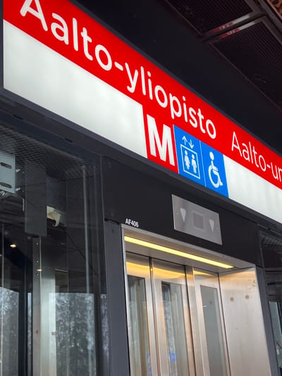 The image displays a modern metro station entrance area at Aalto-yliopisto in Espoo, Finland. A prominent, illuminated overhead sign features a red background with white Finnish text reading "Aalto-yliopisto". Adjacent to this, a red square with a white "M" symbol signifies the metro, and a blue square contains white pictograms for elevator access (two figures with up/down arrows) and wheelchair accessibility. Partially visible text "Aalto-un" extends to the right.

Below the signage, a black horizontal panel sits above a modern elevator entrance. The elevator features stainless steel and glass doors, illuminated by a warm, yellowish LED strip light directly above. On the black panel, to the left of the elevator call panel, the alphanumeric identifier "AF406" is visible. A silver-toned call panel with up and down arrow buttons and a central square indicator is installed directly above the elevator doors. To the left of the elevator, a large glass panel reflects blurred outdoor elements, including bare trees, indicating a connection to an exterior environment. No individuals are present in the scene.