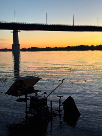 This image depicts a serene waterscape at sunset, likely in Kambarka, Russia, near a large bridge. The sky transitions from a pale blue at the top to a vibrant orange and yellow gradient near the horizon, reflecting on the calm water's surface. A prominent silhouette of a bridge with support pillars and streetlights stretches across the upper portion of the frame. In the foreground, a fishing setup is partially submerged in the water. This setup includes a black feeder seat with various compartments and accessories, such as rod rests, a landing net partially submerged, and a cover. The overall scene conveys a peaceful and tranquil atmosphere, characteristic of an evening by the river.