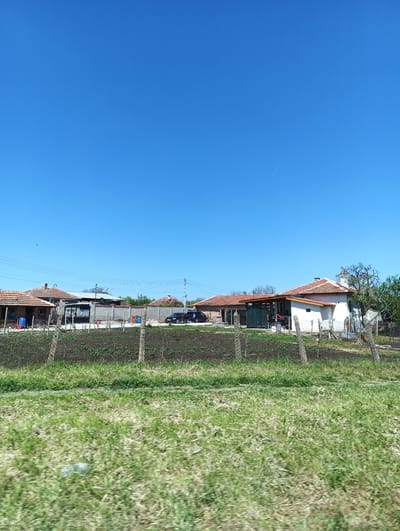 The image depicts a rural scene in Zimnitsa, Bulgaria, under a clear blue sky. In the midground are several residential buildings with red tile roofs, separated by fences. A cultivated garden plot with young plants is visible between the fence and the houses. Parked cars are seen near one of the properties. The foreground consists of a field of green grass. There are no visible people.
