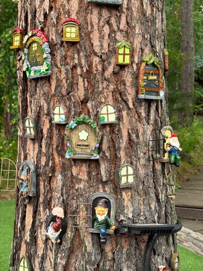 The image depicts a large tree trunk adorned with numerous miniature fairy tale-themed decorations, resembling gnome homes and windows. These decorations are made of resin and are attached to the bark at various heights. Among the houses and windows are several gnome figurines in different poses and attire, some peeking out from windows or perched on ledges. A few of these decorations appear to be suspended by wires or strings. The tree itself has rough, textured bark. The background shows a blurred environment of green foliage and a wooden walkway, suggesting an outdoor setting, possibly a garden or park. The decorations are colorful and whimsical, creating a fantastical scene. The location is explicitly stated as Sukhaya, Russia.