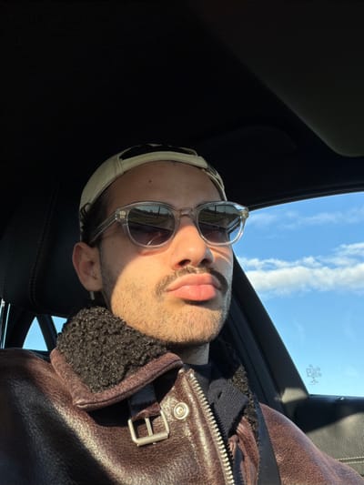 The image displays a male individual seated in the front of a vehicle, in what appears to be the driver's position, located in Enghien, Belgium. The subject's face, characterized by light skin, dark hair, a dark mustache, and a dark stubble beard, is partially illuminated by direct sunlight from the right. He wears clear-framed sunglasses with dark lenses, which reflect a segment of open road and distant vegetation beneath a blue sky. A light-colored baseball cap is worn backward on his head. He is dressed in a dark brown leather jacket with a dark, textured, possibly shearling, collar, over a dark undergarment. A dark seatbelt is secured across his chest. The vehicle's interior features dark upholstery. Outside the vehicle's right-side window, a clear blue sky with scattered white clouds is visible.