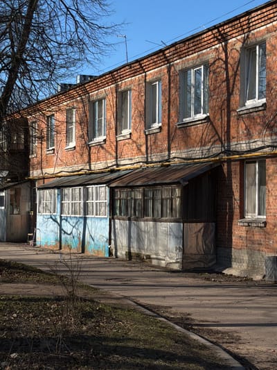 The image depicts a residential building in what appears to be Lavriki, Russia, based on the style and environment. The building has a brick facade, with several windows visible. A covered veranda or extension protrudes from the side of the building, with a mix of materials including wood, metal sheeting, and faded blue paint. The building faces a narrow paved road, with patches of dirt and dried vegetation along the edge. Bare tree branches are visible in the foreground, suggesting it might be late autumn or winter. No people are visible in the frame. The scene is bathed in bright sunlight, casting shadows of the trees and building on the facade. Electrical or utility lines run along the side of the building.
