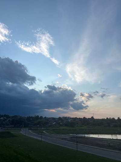 The image depicts a wide-angle view of a landscape that includes a body of water, a road, grassy areas, and a settlement in the background. The sky dominates the upper portion of the image with a mix of dark, heavy clouds on the left and lighter, wispy clouds on the right, suggesting a transition in weather or time of day, possibly near sunset or sunrise. The settlement appears to be comprised of houses and trees, with some structures suggesting a rural or semi-urban environment. A road curves through the middle ground, with streetlights visible along its path. The foreground features a sloped grassy area. The location in Жухавіцкі сельскі Савет, Belarus, can be inferred from the geographical context provided. There are no people or animals clearly visible in the image. The scene is static and appears to capture a moment of natural light and atmospheric conditions over a tranquil setting.