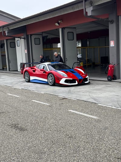 An outdoor scene at the Circuito Tazio Nuvolari in Cervesina, Italy, depicts a red, white, and blue Ferrari 488 GT3 Evo race car parked on a concrete apron in front of a series of dark red garage bays. The car features a large rear wing, multi-spoke silver wheels, and a livery with prominent red sections, white roof and lower body accents, and blue stripes across the front hood and fenders. "FERRARI F488 CHALLENGE" and "OMAR FANTINI CLUB" decals are visible on the windshield. The driver's side door is slightly ajar. Two adult males are positioned behind and to the left of the car's front section, both looking downwards towards the vehicle, indicating inspection or preparation. The garage bays have light grey/white doors, some partially open, revealing yellow safety barriers and equipment within. A red wheeled fire extinguisher is located next to the rightmost visible garage bay entrance. The foreground consists of asphalt pavement marked with two dashed white lines. The sky is overcast, providing diffused illumination.