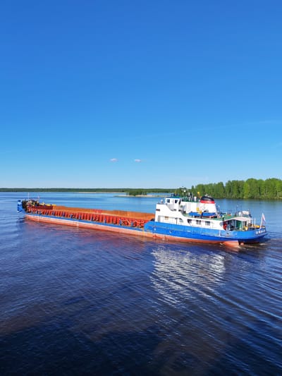 The image presents an elevated, high-angle view of a bulk carrier vessel navigating a broad body of water identified as being near Novodevich'e, Russia. The scene is set under a clear, brilliant blue sky, with a densely forested shoreline forming the distant background.

The vessel is composed of two primary sections: a motorized pusher-tug unit at the stern and a long, open-hold barge segment positioned forward. The pusher-tug features a multi-deck white superstructure atop a blue hull, with a red anti-fouling paint line visible below the waterline. Key features on the tug include a black and red striped funnel, various antennae, radar equipment, and multiple lifeboats. A Russian national flag is affixed to the stern, where the alphanumeric designation "ОК МИ-35" is legible. The barge section, which is significantly longer, has tall, reddish-brown corrugated metal sides. It carries a bulk cargo that appears to be granular material, possibly sand, gravel, or ore, filling its open holds. Blue railings or structural elements run along the upper perimeter of the barge's sides. The entire vessel is in motion, traversing from the mid-left to the mid-right of the frame, leaving a distinct white wake trailing behind the pusher-tug.

The body of water is a deep, dark blue-brown, exhibiting numerous ripples and subtle reflections of the sky and the vessel's hull. The wake created by the moving vessel is prominent, showing lighter, disturbed water patterns.

The sky is uniformly clear and vibrant blue, with minimal cloud cover, consisting of a single faint white contrail or wispy cloud high in the upper-left quadrant and a small, isolated cloud near the center-right.

The distant background features a continuous, dark green band of lush forest, indicative of a wide riverbank or the edge of a large lake. No individuals are visible on the vessel or within the surrounding environment. The scene is illuminated by bright daylight, suggesting a high sun position.