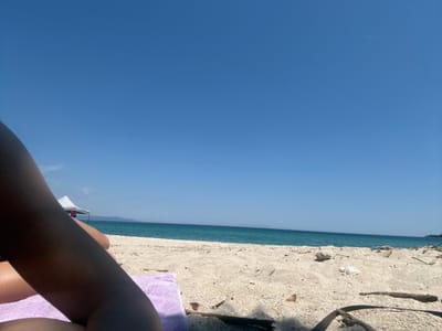 The image depicts a scene at the beach in Ολυμπιάδα, Greece. A clear blue sky occupies most of the frame, contrasting with the narrow strip of turquoise sea along the horizon. In the foreground, there is a sandy beach with visible debris like small twigs and shells. A lavender-colored towel is partially visible on the sand, and a person's legs are positioned in the right part of the frame. A small boat with a white sail is visible on the sea.
