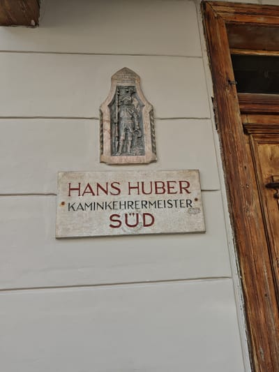 The media file shows a close-up, eye-level view of a sign mounted on a light-colored, horizontally-paneled wall. To the right of the sign is a weathered, dark wooden door frame. Above the sign, a small, relief carving is affixed to the wall, depicting a figure in armor holding a staff. The carving has text above it, though it is difficult to read clearly.
The main subject is a rectangular sign with red lettering. The top line reads "HANS HUBER". Below this, in smaller letters, it says "KAMINKEEHRMEISTER". The bottom line, in larger letters, reads "SÜD". There are two small red dots, one between "HANS HUBER" and "KAMINKEEHRMEISTER", and another to the left of "KAMINKEEHRMEISTER". The sign itself appears to be made of stone or a similar material with a textured surface.
The scene appears to be outdoors, possibly on the facade of a building. The wooden door frame suggests an entrance to a building, and the sign likely identifies a business or resident. The Kitzbühel, Austria location context indicates a European setting. The lighting suggests it is daytime, but the weather is not discernable. The overall impression is of an old-world charm due to the carving and the weathered door.