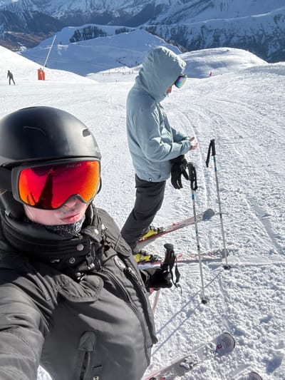 The image displays a snow-covered mountain ski slope in Saint-Sorlin-d'Arves, France, under clear, sunny conditions. Two individuals are equipped for skiing in the foreground and midground, with a third individual visible in the distant background.
The person in the immediate foreground, positioned left, is captured from a slightly elevated, close-up angle, facing the camera as if holding a device. This individual wears a black SINNER brand ski helmet, Oakley brand ski goggles with mirrored red lenses reflecting the snowy surroundings, a black ski jacket featuring multiple zippers and buttons, and a patterned neck gaiter. A black gloved hand is partially visible, holding a ski pole. White Völkl brand skis with yellow and black bindings are partially visible at the bottom right.
To the right and slightly behind the foreground individual stands a second person. This individual is turned away from the camera, facing their hands, appearing to adjust a dark glove on their left hand. They wear a light blue hooded ski jacket with the hood up, dark trousers, and dark ski gloves. Iridescent ski goggles are visible on the side of their head. Red and black Rossignol brand skis with yellow bindings are visible, and two ski poles stand upright in the snow beside them.
In the far left background, a third dark silhouette of an individual, likely a skier or snowboarder, descends the distant slope. Two orange vertical markers are present on the mid-distance slope. The background features extensive snow-capped mountains beneath a bright blue sky. The snow on the slope exhibits texture and tracks from previous use.
