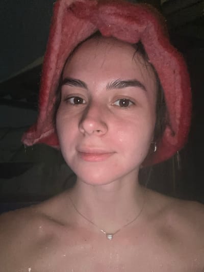 A young adult female is depicted in a close-up, chest-up portrait in the city of Базарово, Russia. She has light skin, dark eyes, and dark eyebrows, with strands of dark hair visible near her forehead. She is wearing a thick, textured, red head covering, possibly a sauna hat, that covers the top and sides of her head. Water droplets are visible on her face, shoulders, and chest, indicating a wet or humid environment. A thin, silver-colored chain necklace with a small, light-colored, geometric pendant hangs around her neck. A small, silver-colored hoop earring is present on her right earlobe. Her skin appears flushed, and she looks directly at the camera with a neutral, slightly smiling expression. The background is dark, out of focus, and indistinct.