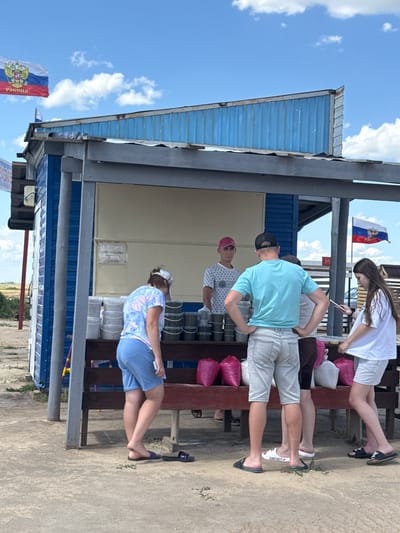 The image shows an outdoor market stall with several people. The stall is a small, blue-colored structure with a wooden counter. There are various items displayed on the counter, including buckets and bags. A Russian flag is visible flying in the background. The ground is made of sand and dirt.
There are at least five people in the image, including a vendor and customers. One person is wearing a light blue t-shirt and light-colored shorts, another is in a white t-shirt and white shorts, and a third is wearing a patterned t-shirt and blue shorts. A fourth person is wearing a red cap and a patterned t-shirt. The fifth person is a young female wearing a white t-shirt and white shorts.
The scene appears to be a casual market setting, possibly in a rural area given the sparse vegetation and open sky. The presence of the Russian flag suggests the location is in Russia. While the exact city of Бурсоль, Russia, is mentioned in the prompt, there are no specific visual cues within the image that definitively confirm this location. The overall atmosphere is that of a warm, sunny day.