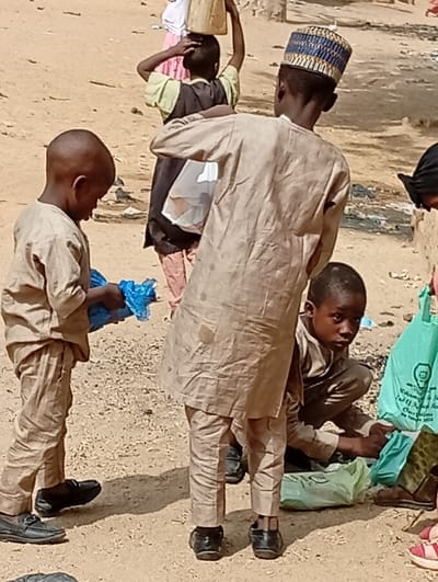 This outdoor image from Fanisau, Nigeria, captures a sunlit scene featuring several children on sandy, dusty ground. The bright daylight suggests it is midday or early afternoon.
In the foreground, a young boy with closely cropped hair stands on the left, facing away from the viewer. He wears a light brown tunic and trousers, with black loafers, and holds a crumpled blue plastic bag. To his right, a taller boy, also dressed in a similar light brown tunic and trousers, faces away and wears a blue and beige knitted cap. He has black shoes.
On the right side of the foreground, a third boy crouches on the ground, actively looking into a light green plastic bag. He is dressed in light brown attire and gazes towards the camera with a focused expression. Another light green plastic bag rests beside him. Visible text on one of these bags includes "Welcome," and partially discernible words such as "Class" or "Club," and possibly a year like "2023" or "2024," along with a circular logo. Partially visible next to the crouching boy is the lower portion of another person, wearing dark clothing and sandals.
In the mid-ground, a child walks away from the viewer, balancing a yellow jerry can on their head. This child wears a light green top, a dark vest, and pink trousers.
The scene depicts children engaged in various activities, possibly involving the distribution or collection of items from the plastic bags, while another child carries water, a common daily task in the region. The setting appears to be a rural or semi-urban area, characterized by its dry, unpaved ground.