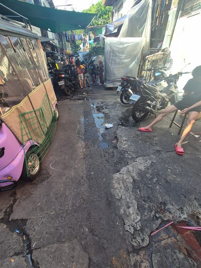 The image shows a narrow street scene in Parañaque, Philippines. The street is paved with asphalt that shows signs of wear and repair, including cracks and patched areas. On the left side of the image, there are buildings, some covered with tarps, and clothing hanging out to dry. A pink motorized tricycle (toktok) with a green rack is parked along the side of the street. Further down the street, people are visible. A person is also visible closer to the foreground, wearing dark shorts and pink sandals, sitting on what appears to be a chair next to a motorcycle. Another motorcycle is parked next to it. The overall impression is of a densely populated, bustling urban environment.