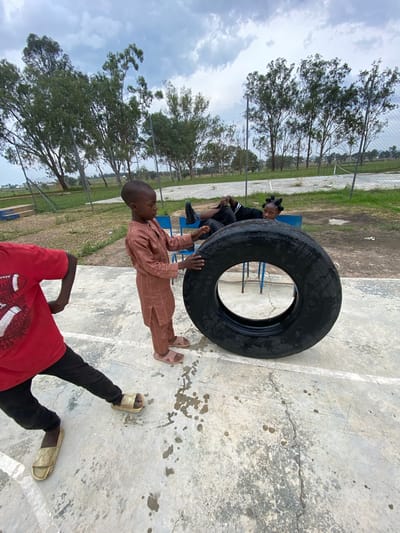 In Zawan, Nigeria, a group of children engage in outdoor activity near a basketball court. A young boy, clad in a brown traditional outfit, stands on a concrete surface, his hands resting on a large, black tire. To his left, another child, wearing a red t-shirt and black pants, is partially visible, their feet adorned with light-colored slides. In the background, two other children are seated, with one reclining and the other looking towards the viewer. The scene unfolds under a cloudy sky, with trees and a fence framing the area.