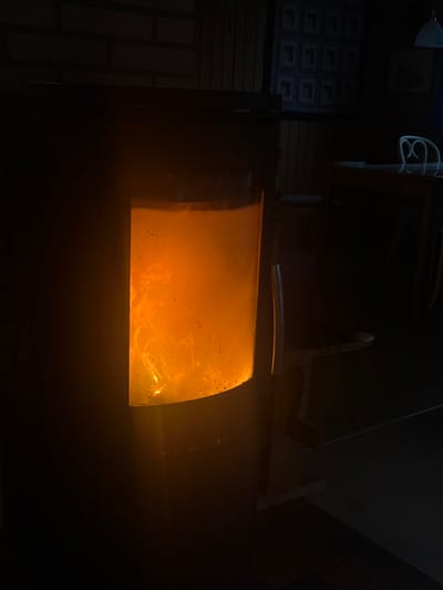 The image shows a close-up of a wood-burning stove with a fire inside. The fire is burning brightly, illuminating the glass door of the stove. The flames are orange and yellow, and there is some smoke visible. The stove is in a dark room, and there is a brick wall visible in the background. The location is in Blåvand, Denmark, at the coordinates 55.555, 8.14.  A wooden table with a white object on top is in the background, slightly out of focus. The scene is a dark interior with the stove being the only source of light.