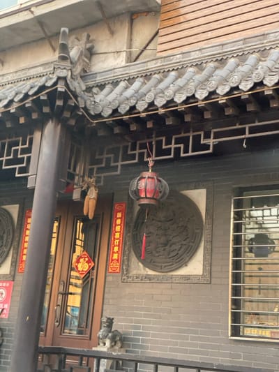 The image displays a detailed street-level exterior view of a building in Taihe District, China, exhibiting traditional Chinese architectural elements and festive decorations.

The building features a dark gray tiled roof with ornate eaves, including a decorative finial on the top left corner. Below the eaves, a dark-colored lattice pattern is visible. The facade is constructed from dark gray bricks. A prominent dark pillar stands on the left, supporting the overhanging roof structure.

A red and dark-colored Chinese lantern, adorned with a red tassel, hangs from the eaves towards the right. To the left of the main entrance, two dried corn cobs are suspended. The entrance consists of a double wooden door with multiple glass panels, reflecting ambient light. Affixed to one of the door's glass panels is a red diamond-shaped paper decoration featuring the inverted Chinese character "福" (fú). The door is flanked by two vertical red banners with gold Chinese characters; the right banner reads "新春喜订菓".

Mounted on the brick wall are two large circular bas-reliefs. The one on the right is clearly visible, depicting a complex design of what appears to be a mythical creature, possibly a dragon, within a decorative circular frame. To the far right, a window with horizontal metal security bars is present.

In front of the building, a dark-colored metal railing runs across the lower portion of the frame. Two small, gray stone statues, resembling guardian lions or Foo Dogs, are positioned on a ledge behind the railing. A small, partially obscured red and white sign with a "no" symbol is visible on the far left. No individuals are present in the scene.