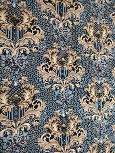 This image depicts a close-up view of a fabric with an ornate, damask-inspired pattern. The pattern features stylized floral elements in shades of gold and beige, intertwined with darker black and grey motifs. A repeating geometric background in muted teal and grey hues forms the base of the design. Within the floral arrangements, there are black diamond shapes containing a gold "S" logo topped with a small crown. Small clusters of blue and grey flowers are also present throughout the pattern. The lighting appears to be even, suggesting an indoor setting or overcast conditions. There is no indication of the city of Pitnak, Uzbekistan, or any specific environmental context within this image.