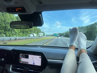 The image shows the interior of a car looking out through the windshield at a highway. In the foreground, a person's legs and feet are resting on the dashboard. The person is wearing white socks with green trim and light-colored ribbed pants. The car's infotainment screen is visible on the right side of the dashboard, displaying a navigation map. The map indicates the location is near Yulee, Florida, United States, with the route showing I-95 North towards an airport in Florence, South Carolina. Outside the car, trees line the highway, and several vehicles, including trucks and cars, are visible on the road. The sky is blue with some clouds. A "PASSENGER AIR BAG ON" indicator light is visible on the overhead console.
