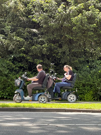 Two people are riding mobility scooters on a sidewalk, next to a road in Dronten, Netherlands. The woman in front is wearing a dark shirt, blue jeans, and glasses, with a backpack. The man behind her is wearing a dark shirt and tan pants, also with a backpack. They are driving past trees and green shrubbery. The road is to their right, with patches of shadows falling across its surface.