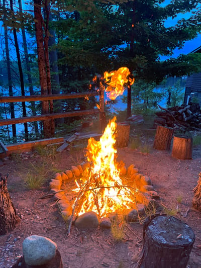 This is a medium shot, eye-level, outdoor photograph taken at dusk in Brantingham, United States. The focal point of the image is a roaring bonfire in a circular fire pit made of rocks. The flames are bright orange and yellow, with sparks rising into the twilight sky. Surrounding the fire pit are several tree stumps, which appear to be used as seating. In the background, there is a wooden fence and a body of water, likely a lake, bordered by tall trees. The overall mood of the image is tranquil and inviting, evoking a sense of peace and relaxation. The location appears to be a rustic, natural setting, possibly a campsite or a backyard with a view of the lake. The sky is a deep blue, indicating that the sun has set, and the trees are silhouetted against the darkening sky. The presence of fire and the natural surroundings suggest an outdoor gathering or a peaceful evening spent by the fire.