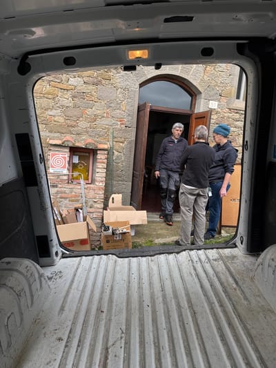 The image presents a view from the interior of a white cargo van, looking out onto an outdoor scene in what is identified as Cortona, Italy. The van's ribbed white floor and clean white interior walls are visible in the foreground, with an illuminated ceiling light fixture above the rear opening.

Three individuals are standing outdoors, engaged in conversation, in front of an ancient stone building.
*   The leftmost person is a middle-aged male with grey hair and a dark beard, wearing a dark work jacket with red accents, grey work pants with black knee pads, and dark shoes with orange soles. He stands facing the other two.
*   The central person, seen from the back, has short light hair, wears a black long-sleeved top, light brown pants, and dark shoes. A lanyard with an ID card hangs from their right side. This person is directly facing the leftmost individual.
*   The rightmost person wears a blue knit beanie, a dark long-sleeved top, blue jeans, and dark shoes. This individual is also facing the leftmost person, with their back slightly angled toward the camera.

The background features a rustic building constructed of irregular grey and brown stone blocks. A large, dark brown wooden double door with an arched top is partially open, revealing a dark interior. A white house number "55" is affixed above the doorway. To the left of the door, a small rectangular window with a red-tiled overhang is set into the stone wall. Below this window, a white sign with a red coiled hose emblem and the Italian word "IDRANTE" (Hydrant) is mounted. A yellow sign with the text "CRASIE" is visible inside the window frame.

On the ground in front of the building, several open cardboard boxes are present. One large box, marked "RGS," contains miscellaneous items. Another box holds various small objects and hardware. A pane of clear material, possibly glass or plastic, stands upright, leaning against the stone wall between the window and the doorway. The ground is a mix of dirt and sparse green vegetation. The sky appears overcast.
