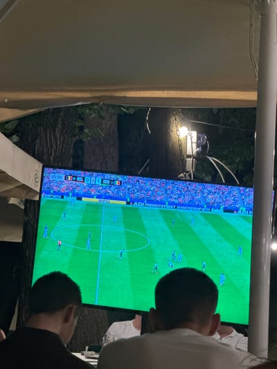 The image depicts a scene in Suceava, Romania, where individuals are gathered to watch a soccer match on a large outdoor screen. The screen displays a game with the score showing Belgium (BEL) with 0 and Senegal (SEN) with 1. The time displayed is 45:59. The soccer field is vividly green, with players in blue uniforms on the screen. In the foreground, two men are visible from behind, engrossed in watching the match. One is wearing a black shirt, and the other, further back, is wearing a white shirt. The setting appears to be an outdoor seating area, possibly a restaurant or bar, with a canopy overhead and foliage in the background, suggesting an evening or nighttime event. A lamp hangs from a chain, illuminating the area. The overall atmosphere suggests a shared viewing experience of a sporting event.