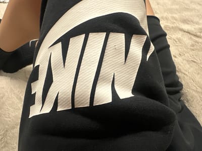 A close-up image shows a person resting, wearing black athletic wear featuring a large white "NIKE" logo. The logo, complete with its characteristic swoosh, is prominently displayed across the fabric, which appears to be a black cotton or fleece material, possibly part of sweatpants or a sweatshirt. Subtle textural patterns are visible within the white letters of the logo and on the black fabric. A small, dark speck is visible on the letter "I." To the left of the garment, a segment of a bare human leg with light-to-medium skin tone is visible, ending in a foot and ankle covered by a plain black sock. The background consists of a light, neutral-colored, plush, and textured material, resembling a shag carpet or a soft blanket, suggesting an indoor setting.